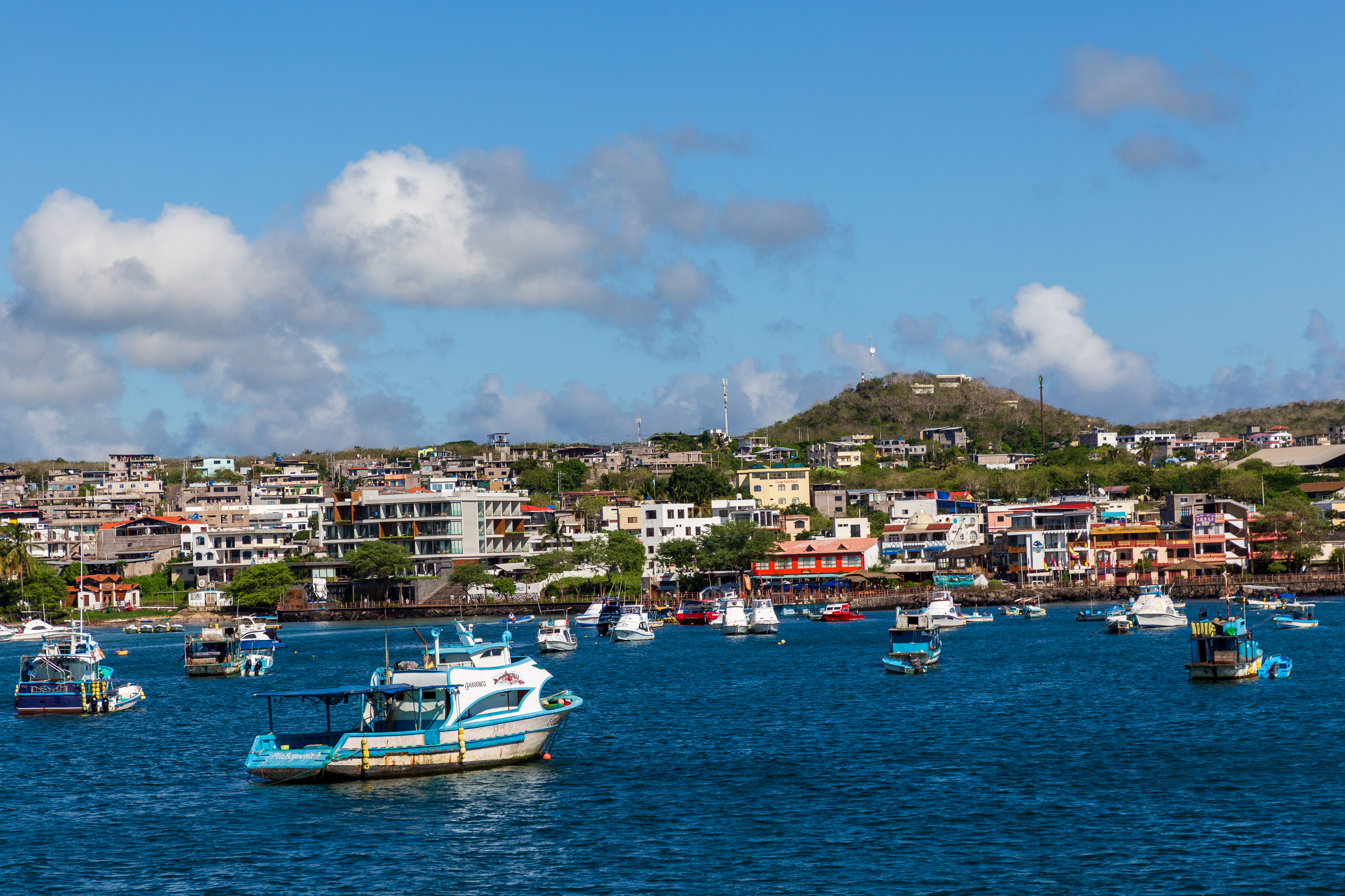 Wide view of the harbor town with boats anchored in deep blue water under a partly cloudy sky